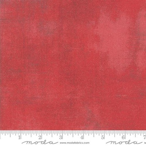 May include: A red fabric with a mottled, textured appearance featuring subtle variations in tone and depth. A white measuring tape is positioned along the bottom edge, showing markings in inches from 0 to 7, with the text moda and modafabrics.com.