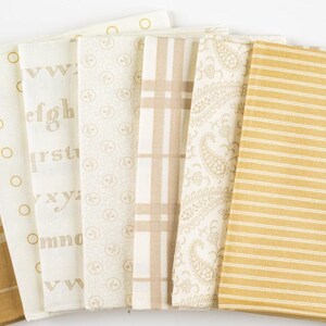 May include: A collection of six folded fabric swatches in shades of cream, beige, and mustard yellow. The patterns include a cream fabric with gold alphabet letters, a beige circular motif, a beige plaid, a cream paisley, and a mustard striped fabric.