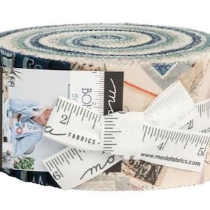 May include: A rolled bundle of fabric with a mix of blue, cream, and tan colors. The fabric is wrapped with a white measuring tape and a tag that reads "moda FABRICS + SUPPLIES". The fabric roll is approximately 6 inches in diameter.