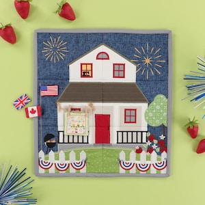 May include: A square, quilted wall hanging featuring a white house with a red door and windows, a brown roof, and a picket fence. The background is blue with embroidered fireworks. Small flags and strawberries surround the house.
