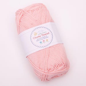 May include: A skein of light pink chunky cotton yarn wrapped in a white paper label. The label features a scalloped circular border and text that reads 100% Cotton Chunky Thread by Lori Holt of Bee in my Bonnet, with small flower illustrations.
