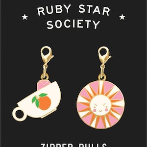 May include: Two gold zipper pulls with charms. One charm is a white teacup with a pink rim and a peach inside. The other charm is a pink sun with a yellow face and orange rays. The text "RUBY STAR SOCIETY" and "ZIPPER PULLS by Melody Miller" is printed below the charms.