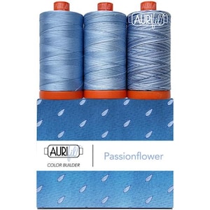 May include: Three spools of blue sewing thread with orange bases, arranged in a row. The spools feature the Aurifil logo on the top and side. They are positioned above a blue rectangular box labeled Aurifil Color Builder Passionflower.