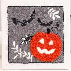 May include: A Halloween-themed, square, gray and white stitched artwork. The design features a smiling orange pumpkin, two black bats with floral patterns, and white leaf sprigs. The artwork is framed with a black border.