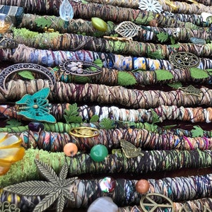 May include: An assortment of handmade dreadlock wraps in various colors and patterns, adorned with decorative charms, beads, and leaves. The wraps are arranged on a wooden surface with moss and small stones, creating a natural, bohemian aesthetic.