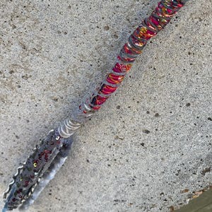 May include: A long, slender wand-like object wrapped in a vibrant, multi-colored fabric featuring shades of pink, orange, and gold. The handle section is adorned with grey lace, silver sequins, and small metallic beads, creating a textured finish.