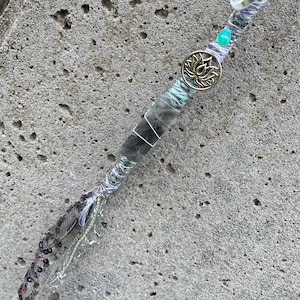 May include: A handcrafted hair wrap featuring a gray stone wrapped with silver wire and adorned with teal beads, a gold lotus charm, and iridescent accents. The wrap is approximately 8 inches long.