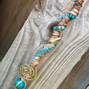 May include: A hair accessory featuring a long, flexible cord wrapped in tan and brown fibers, accented with small turquoise beads. It includes a gold-toned metal spiral charm and a single large, round turquoise bead at the bottom end.