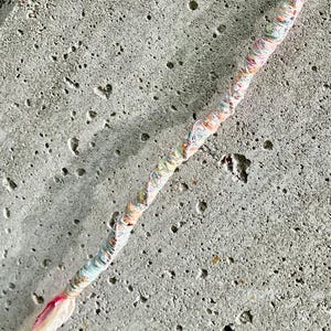 May include: A hair wrap featuring a multi-colored thread design with pastel shades of pink, blue, yellow, and green tightly wound around a central core, ending in loose, flowing strands of pink and white thread at the bottom tip of the accessory.