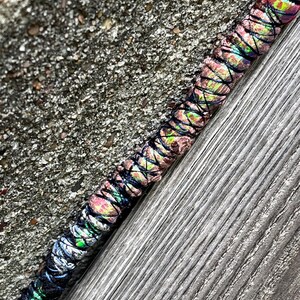 May include: A colorful dreadlock wrap with a vibrant pattern of pink, green, and blue hues. The wrap is intricately woven with black thread, creating a textured appearance. The wrap is resting on a weathered wooden surface.