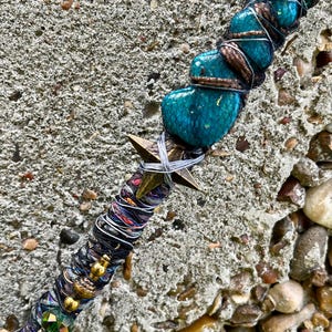 May include: A handcrafted wand featuring turquoise-colored stones, wrapped with silver wire and brown cord. A bronze star and decorative beads adorn the wand's center and base. The wand is set against a textured, gray background.