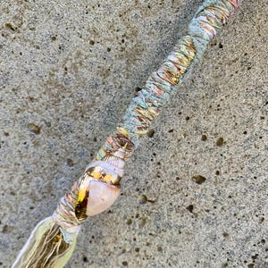 May include: A decorative wand wrapped in strips of fabric in shades of sage green, pale pink, and metallic gold. A small, iridescent seashell with gold-toned accents is attached near the bottom, which ends in a frayed, light-colored fabric tassel.