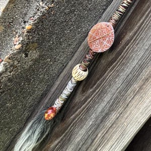 May include: A handcrafted hair stick with a decorative design. The stick features a variety of beads in silver, brown, and cream colors, along with a patterned oval pendant. The end of the stick has a crystal-like embellishment and a wispy, light-colored accent.