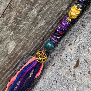 May include: A handcrafted hair wrap featuring a gold owl charm, a green bead, and a gold pentagram charm. The wrap is adorned with purple, blue, and pink threads, and a tassel of similar colors. The background is a weathered wooden surface.