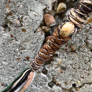 May include: Close-up of a handmade, wrapped item with a natural, earthy aesthetic. The main body is wrapped in brown and beige twine, with a lighter-colored bead. The end features strands of green, black, white, and tan yarn.