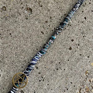 May include: A hair wrap featuring a long, thin cord wrapped in various shades of black, white, and teal thread. A circular brass-toned metal pentagram charm is attached to the lower section of the wrap, secured by the winding thread pattern.