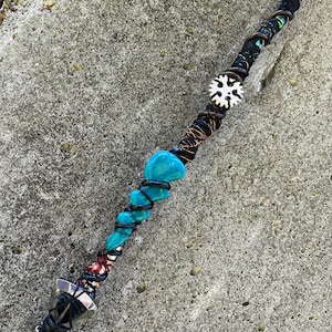 May include: A handcrafted wand with a black handle wrapped in fabric and wire. Turquoise stones are wrapped in black wire along the wand's length. A white snowflake-shaped charm and a silver-colored tip complete the design.
