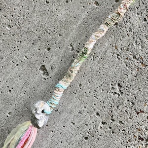 May include: A hair wrap featuring a long, thin cord wrapped in various pastel-colored threads including green, blue, and cream. A small, grey, textured stone bead is attached near the end, which features a tassel of frayed fabric strips.