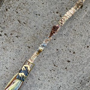 May include: A hair wrap featuring a rectangular charm with a blue and white wave design, a small gold-toned fish charm, and a brown spiral shell charm. The wrap is constructed from multi-colored fabric strips in shades of blue, yellow, and cream.