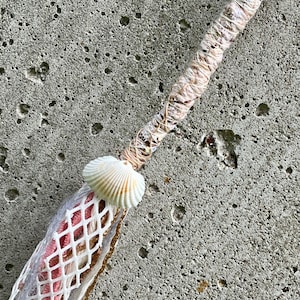 May include: A decorative wand featuring a long handle wrapped in light-colored twine. A white ridged seashell is attached at the base of the handle, which transitions into a tassel made of white netting, pink fabric strips, and gold-toned fibers.