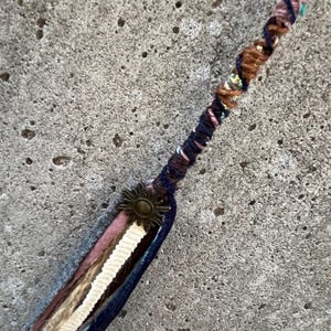 May include: A handmade hair wrap featuring a sun-shaped metal accent. The wrap incorporates various fabrics in shades of blue, pink, brown, and cream. The design includes twisted strands and decorative ribbons, creating a bohemian aesthetic.