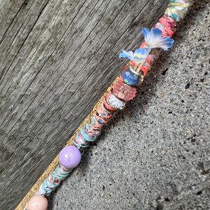 May include: A decorative wand with a colorful, textured design. The wand features a variety of embellishments, including pink, blue, and purple beads, and small blue flower accents. The wand is wrapped in colorful fabric.