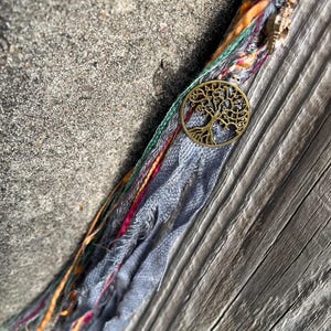 May include: A handmade hair wrap featuring a bronze tree of life charm. The wrap includes strands of gray, orange, green, and pink fabric. A small gold-colored charm and a light blue bead are also visible.