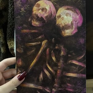 Puede incluir: Una pintura de dos cr&aacute;neos humanos con una figura esquel&eacute;tica en el fondo. Los cr&aacute;neos est&aacute;n pintados en tonos de rosa y amarillo, y la figura esquel&eacute;tica est&aacute; pintada en tonos de marr&oacute;n y dorado. El fondo es de color morado oscuro.