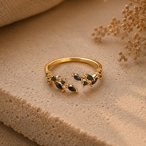 May include: A gold-toned open ring featuring two symmetrical branches adorned with small, marquise-cut black stones. The band is adjustable, with the two ends meeting in the centre, each decorated with a cluster of dark, leaf-shaped gemstones.