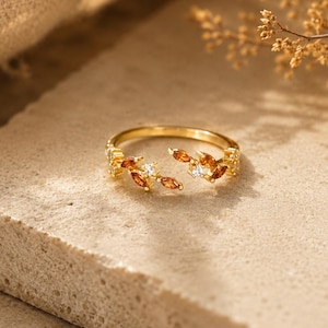 May include: A gold ring with a leaf design, featuring orange-brown gemstones and clear accents. The ring is set on a textured, neutral surface. The design is delicate and detailed, showcasing the ring's craftsmanship.