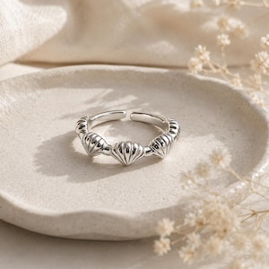 Peut inclure: Une bague ouverte de couleur argentée ornée d'une série de motifs de coquillages en relief répétés tout autour de l'anneau. La bague présente une finition métallique polie et une ouverture réglable à l'arrière pour s'adapter aux doigts.