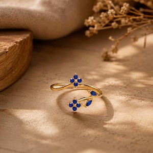 May include: A gold-toned open ring featuring two floral motifs composed of four small round blue stones each, accented by two marquise-cut blue stones along the band. The ring has a bypass design with a polished metal finish and vibrant blue gems.