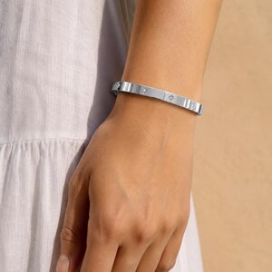 Peut inclure: Un bracelet jonc en métal argenté orné d'une série de petits motifs en forme d'étoiles gravés et espacés régulièrement sur toute la bande. Le bracelet présente une finition lisse et polie ainsi qu'un profil plat et uniforme autour du poignet.
