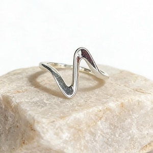 May include: A silver-coloured ring featuring a single, continuous wave-like design that rises to a central peak. The band is smooth and reflective, showcasing a metallic finish that curves into a distinct zigzag shape across the top of the ring.
