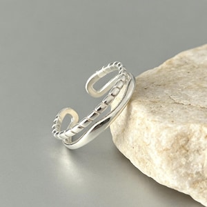 Peut inclure: Une bague ouverte de couleur argentée présentant un design à double anneau. Un anneau possède une finition lisse et polie, tandis que l'autre affiche un motif texturé rappelant une corde. La bague est ajustable avec une ouverture à l'arrière.