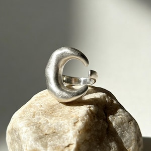 May include: A silver-toned open ring featuring a crescent moon shape with a brushed, matte finish. The band is adjustable with a gap at the back, showcasing a smooth, curved design that tapers at the ends, creating a distinct celestial aesthetic.
