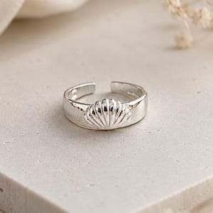 May include: A polished silver-tone ring featuring a prominent, raised seashell motif centred on the band. The ring has an open-back design, allowing for adjustable sizing, with a smooth, reflective metallic finish across the entire surface.