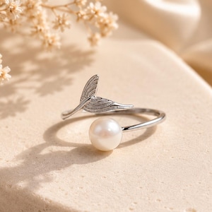 Peut inclure: Une bague ouverte de couleur argentée dotée d'une perle blanche polie à une extrémité et d'un motif texturé en forme de nageoire à l'autre. L'anneau est fin et métallique, créant un style ouvert permettant un ajustement réglable.