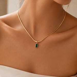 May include: A gold-toned snake chain necklace featuring a small, rectangular pendant with a deep green stone set in a gold-coloured frame. The necklace rests against the collarbone, showcasing a smooth, metallic finish and a simple, refined design.