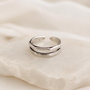 May include: A silver-toned open ring featuring a double-band design. The top band has a subtle hexagonal shape, while the bottom band is smooth and rounded. The ring is adjustable with a small gap at the back, showcasing a polished metallic finish.