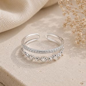 May include: A silver-toned open ring featuring two bands. The top band is encrusted with small clear stones, while the bottom band displays a repeating pattern of small shell-shaped motifs alternating with individual clear stones.