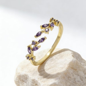 May include: A gold-colored ring with a delicate design. The ring features a series of marquise-cut purple gemstones arranged in a leaf-like pattern. The ring is set against a white background, highlighting its intricate details and elegant style.