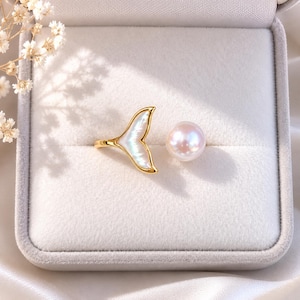 Può includere: Un anello aperto color oro con un ciondolo a forma di coda di balena in madreperla bianca iridescente su un'estremità e una singola perla bianca rotonda sull'altra, presentato all'interno di un portagioie foderato in velluto grigio chiaro.
