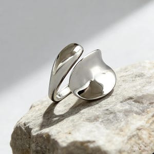 May include: A polished silver ring with a contemporary design. The ring has a teardrop-shaped feature and a curved band. The reflective metal surface creates a shiny effect. The ring is presented on a light-coloured, textured surface.