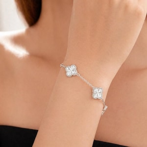 Peut inclure: Un bracelet argenté orné de motifs de trèfles à quatre feuilles. Chaque trèfle est serti de petites pierres scintillantes. Le bracelet est porté au poignet, avec la chaîne et les motifs visibles.