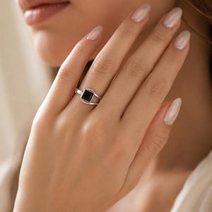 May include: A silver ring featuring a square-cut black stone set in a polished metal band. The band has a split design that converges at the setting, holding the dark, opaque square gemstone securely in the centre of the silver frame.