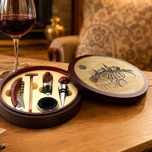 May include: A round wooden wine accessory set case with a dark brown exterior and a light wood lid featuring a black etched vineyard illustration. The interior contains a corkscrew, a thermometer, a bottle stopper, and a circular black drip ring.