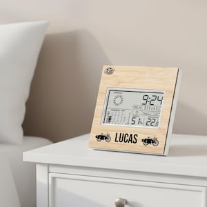 Puede incluir: Una estación meteorológica digital con marco de madera clara muestra la hora, el pronóstico del tiempo y la temperatura. El nombre "LUCAS" está impreso en negro, con gráficos de motocicletas. La temperatura es de 22,8 grados Celsius.