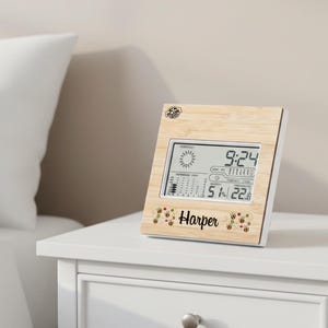 Puede incluir: Estación meteorológica digital con marco de madera clara, mostrando la hora 9:24, una temperatura de 22,8°C y una humedad del 51%. El nombre "Harper" está impreso debajo, con gráficos de abejas y flores.