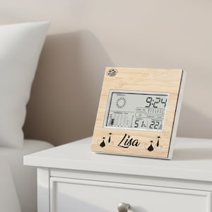 Puede incluir: Una estación meteorológica digital con marco de madera clara, que muestra la hora 9:24. La pantalla muestra pronósticos del tiempo, humedad al 51% y temperatura a 22,8 grados Celsius. El nombre "Lisa" está impreso debajo, con siluetas de gatos.